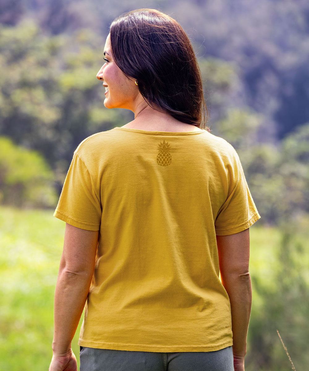 A woman viewed from behind wearing a mustard yellow short-sleeve t-shirt with a small pineapple graphic on the upper back, set against a blurred green outdoor background. View 3