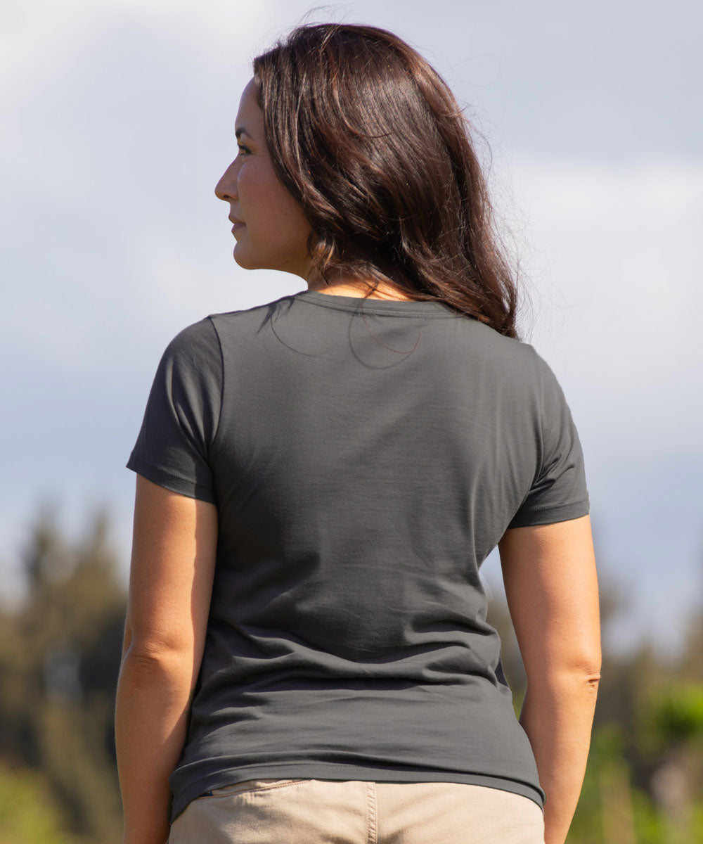 A woman viewed from behind wearing a dark gray short-sleeve crewneck t-shirt and light beige pants, standing outdoors against a soft-focus sky and trees. View 3