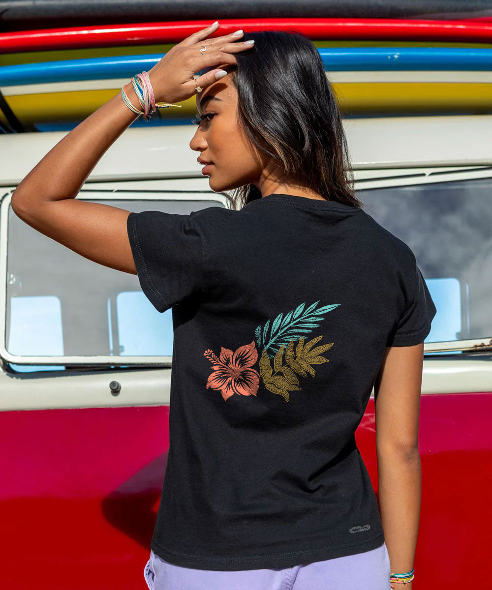A woman wearing a black t-shirt with a colorful tropical floral and leaf graphic on the back, standing in front of a red and white vehicle. View 3
