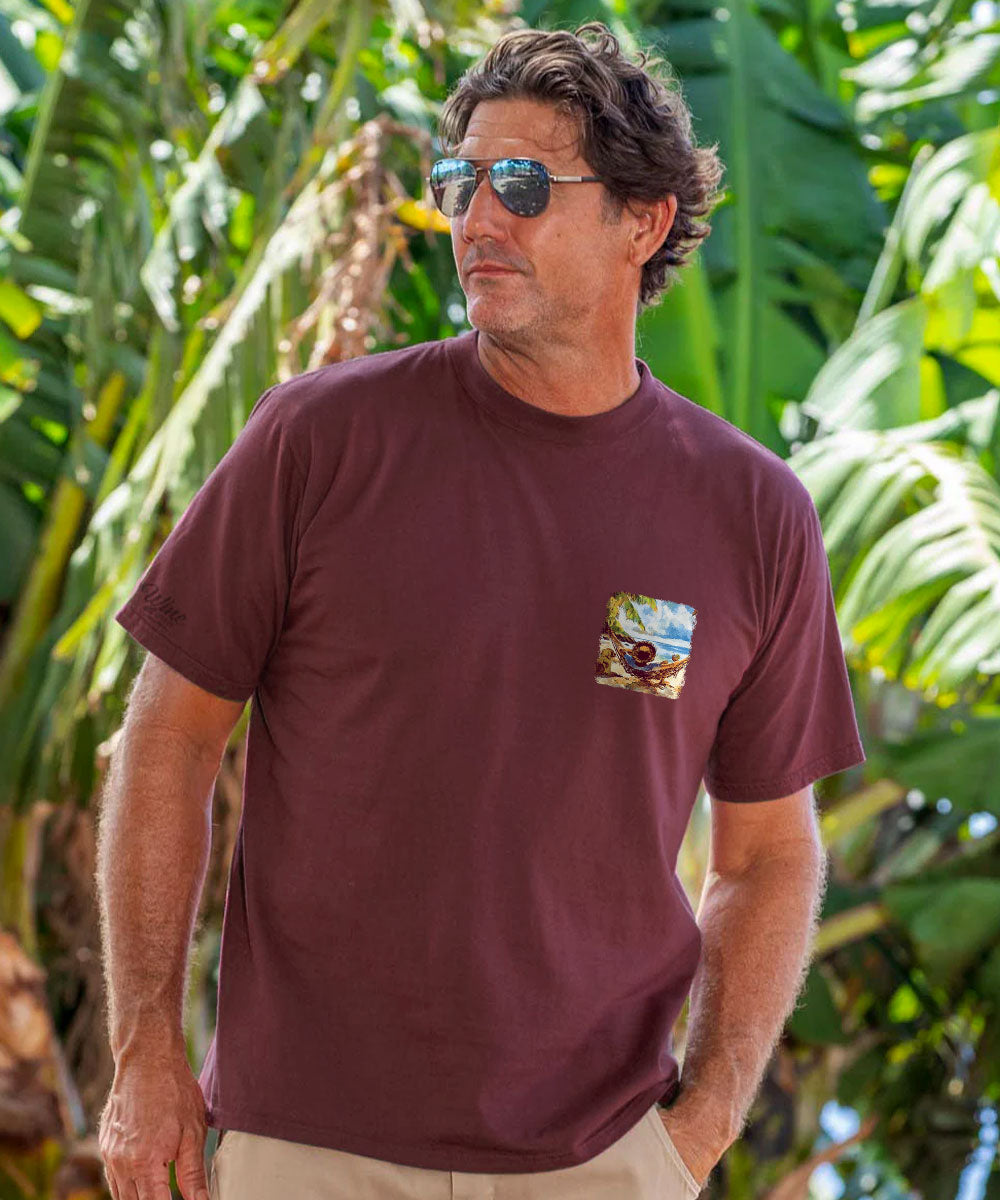 A man wearing a short-sleeve crewneck t-shirt in maroon with a small landscape graphic on the left chest, set against a background of lush green tropical leaves. View 3