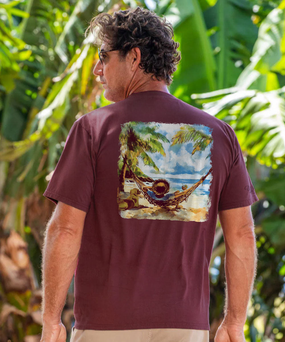 A man wearing a maroon short-sleeve t-shirt with a square graphic print on the back showing a tropical beach scene with a hammock, photographed outdoors against a background of green tropical leaves. View 2