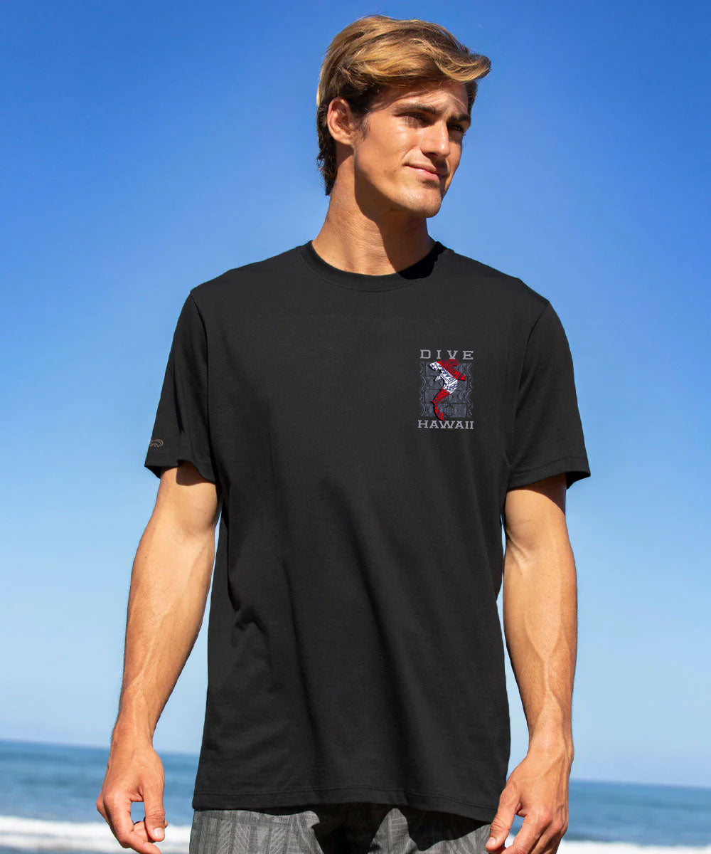 A man wearing a black short-sleeve t-shirt with a small red, white, and blue graphic on the left chest, standing against a bright blue sky and ocean background. View 3