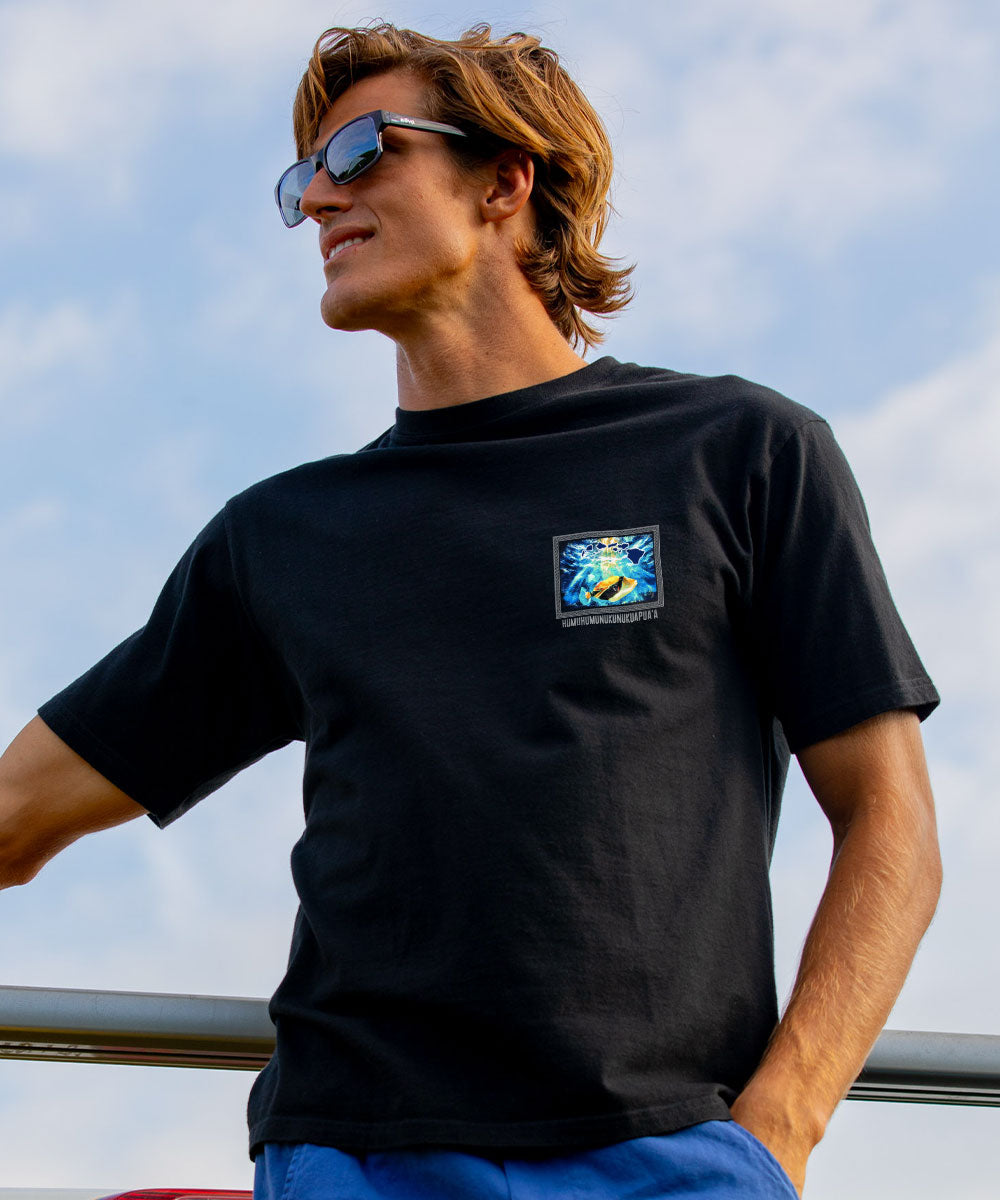 A man wearing a black crewneck t-shirt with a small square graphic on the left chest, photographed outdoors against a cloudy blue sky. View 3