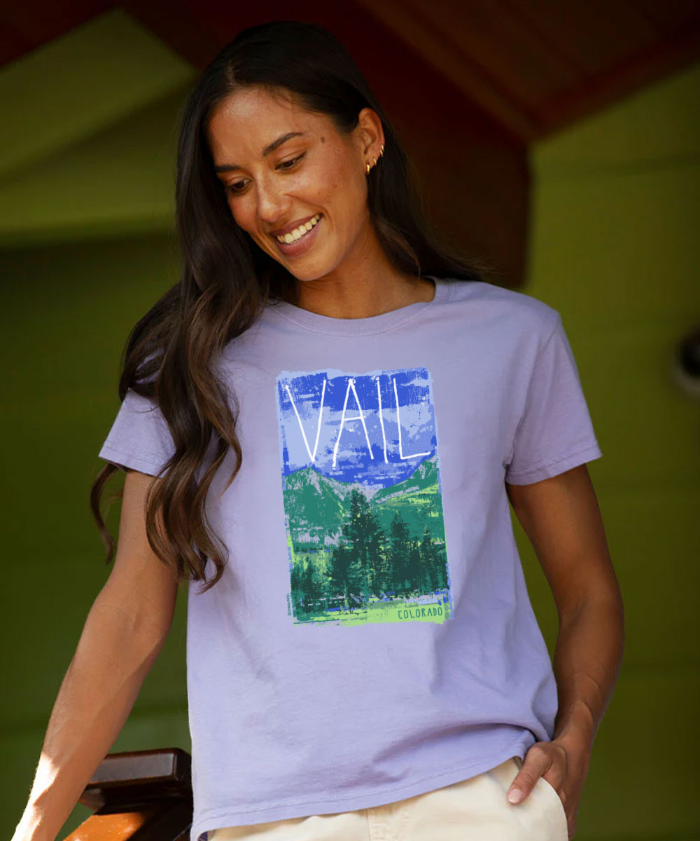 A woman wearing a light lavender graphic t-shirt with a colorful mountain landscape print and the word 'VALE' on the front, paired with cream pants, set against a green outdoor background. View 2