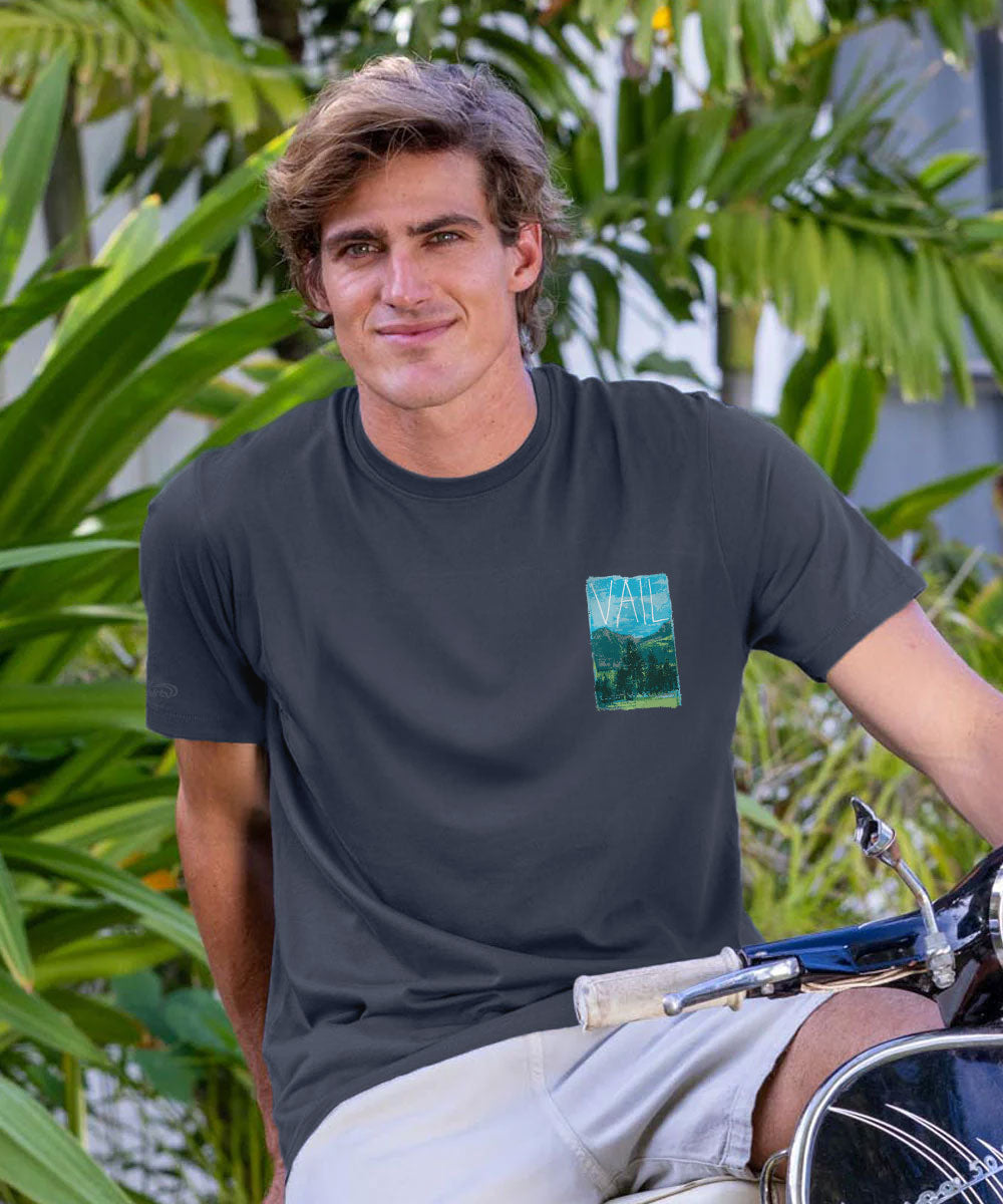A man wearing a dark charcoal gray t-shirt with a small green landscape graphic on the left chest, posing on a motorcycle in front of lush green tropical foliage. View 3