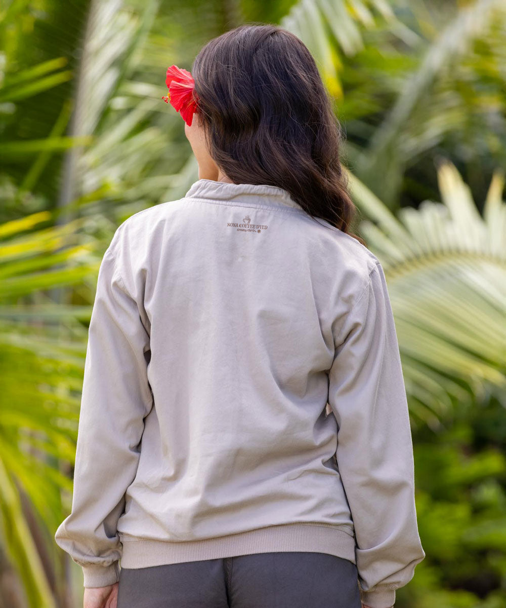 A woman viewed from behind wearing a light beige long-sleeve zip-up jacket, with a red flower in her hair, set against a background of green tropical palm leaves. View 3