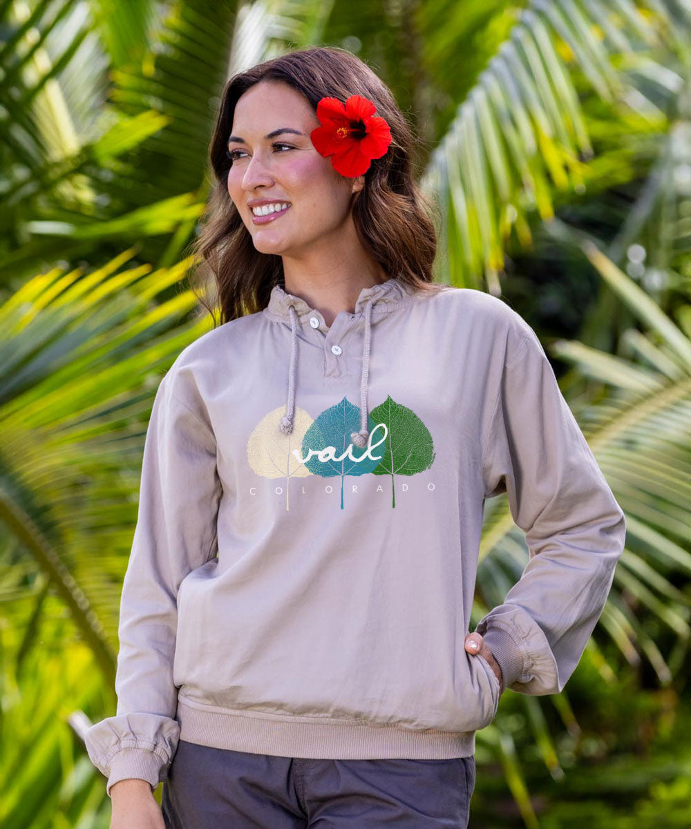 A woman wearing a light taupe pullover hoodie with a 'rail COLORADO' graphic printed on the chest, posed outdoors against a background of green palm leaves. View 2