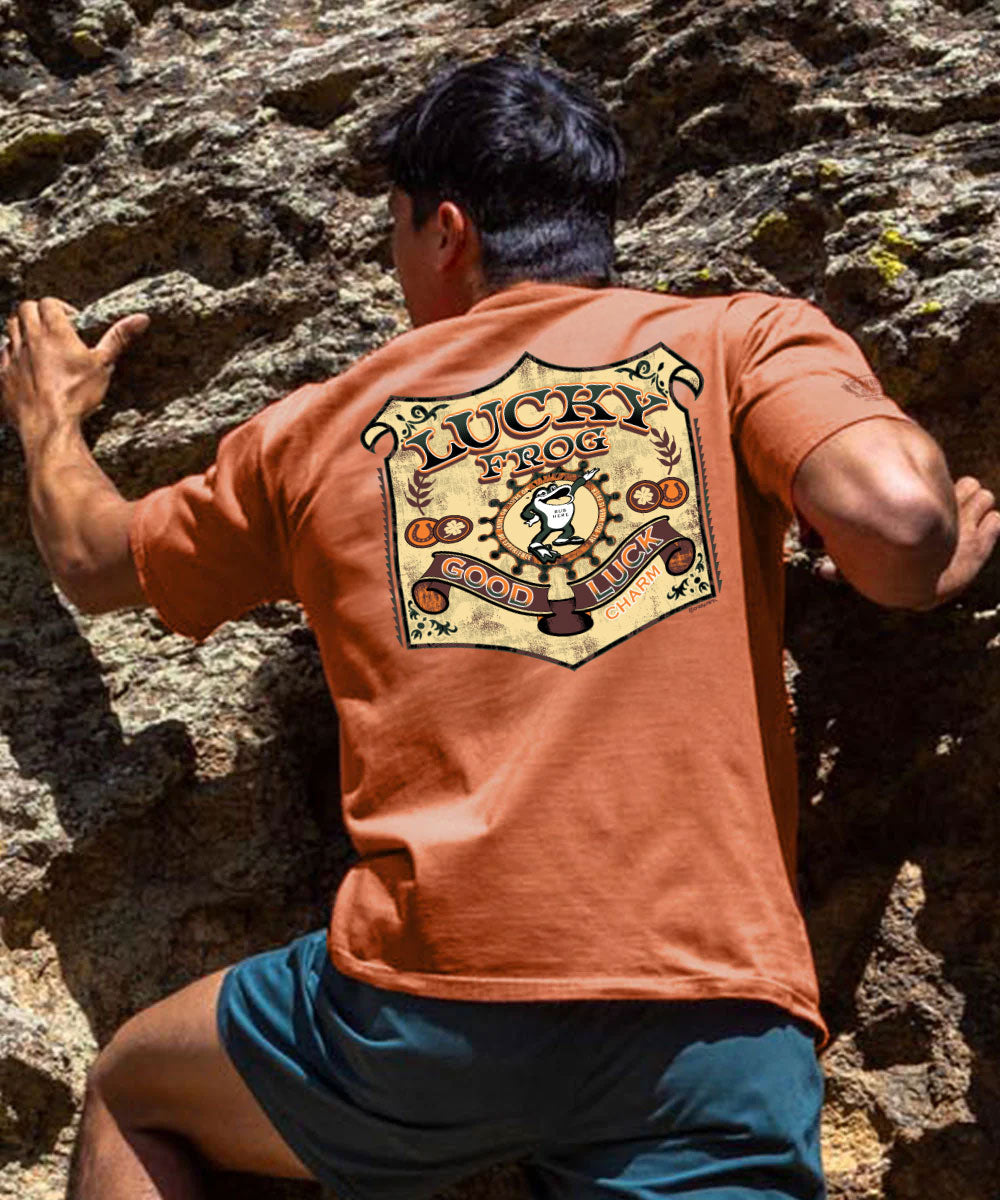 A man climbing a rocky wall, viewed from behind, wearing a burnt orange t-shirt with a large 'Lucky Dog' graphic on the back and teal shorts. View 2