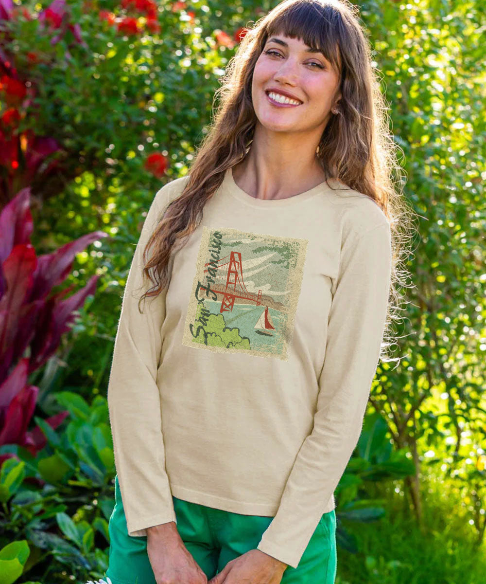 A woman wearing a beige long-sleeve t-shirt with a vintage-style San Francisco Golden Gate Bridge graphic, paired with green pants, standing in a lush garden setting. View 2