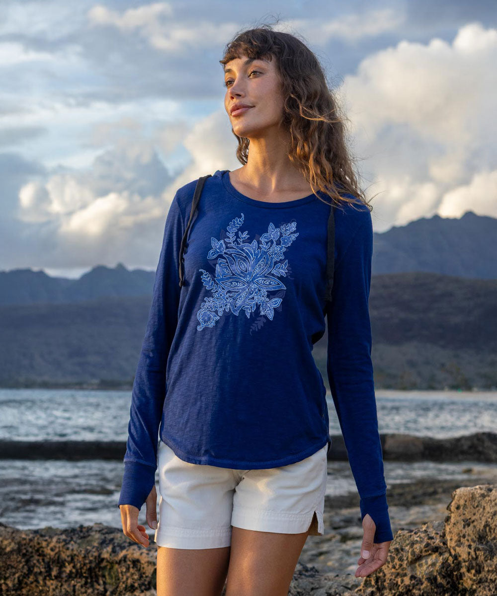 A woman wearing a royal blue long-sleeve t-shirt with a light blue floral graphic on the front, paired with white shorts, standing on a rocky shoreline with mountains and water in the background. View 2