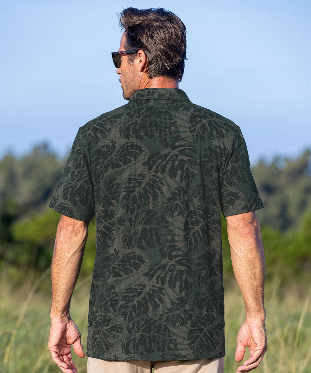 A man wearing a dark green short-sleeve button-up shirt with a tonal leaf pattern, shown from the back in an outdoor setting with a blurred background of trees and sky. View 3
