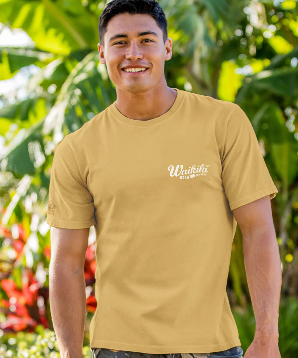 A smiling man wearing a tan crewneck t-shirt with a white 'Wadikiti' logo on the left chest, set against a lush tropical green background. View 3
