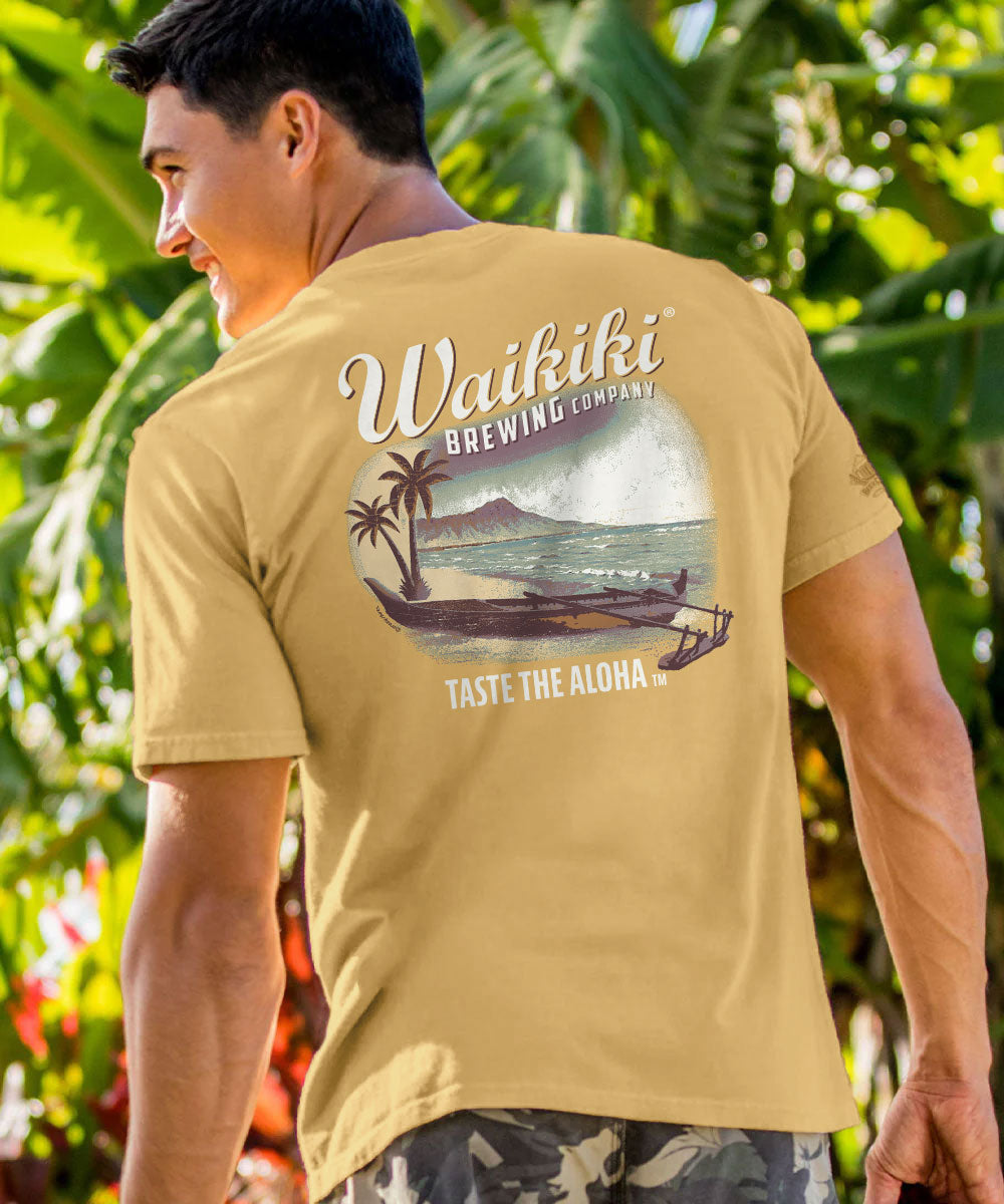 A man wearing a tan short-sleeve t-shirt with a large back graphic that reads 'Waikiki Brewing Company' and 'Taste the Aloha,' standing outdoors in front of lush green tropical foliage. View 2