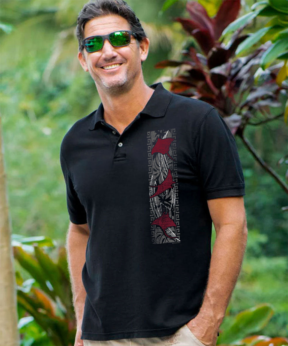 A man wearing a black short-sleeve polo shirt with a vertical graphic print on the chest, standing outdoors in front of a lush green tropical background. View 2