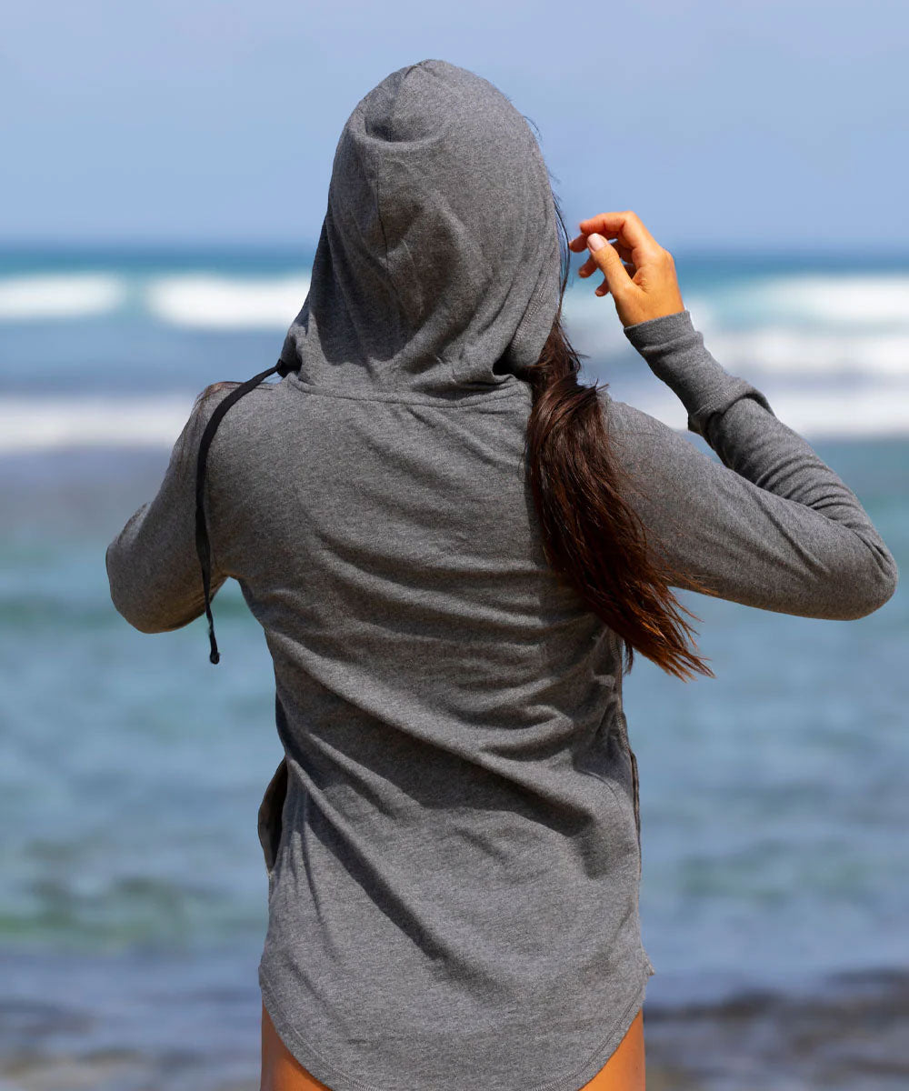 A woman viewed from behind wearing a heather gray long-sleeve hooded shirt on a beach with the ocean in the background. View 3
