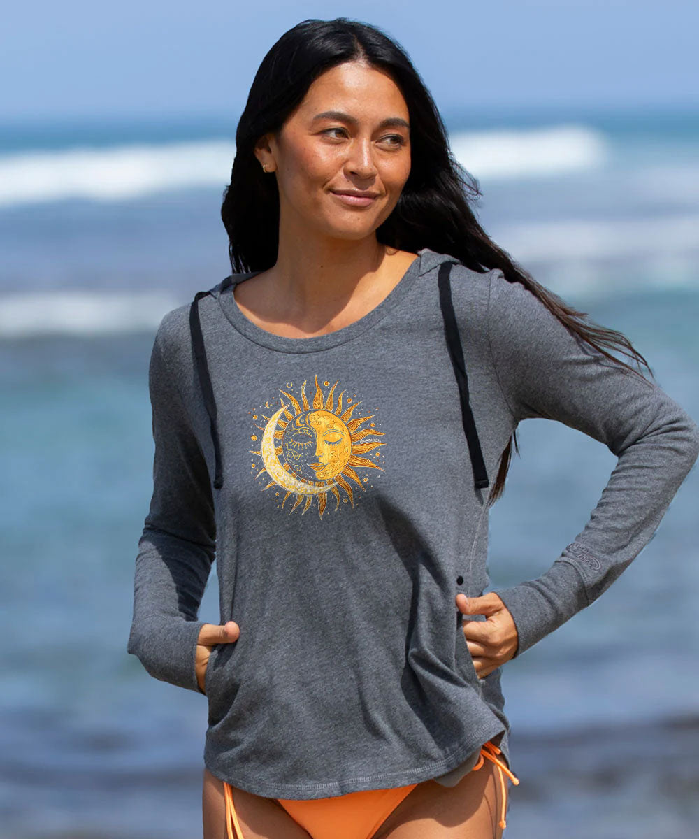 A woman standing on a beach wearing a heather gray long-sleeve top with a yellow sun and crescent moon graphic on the front, paired with orange bikini bottoms.