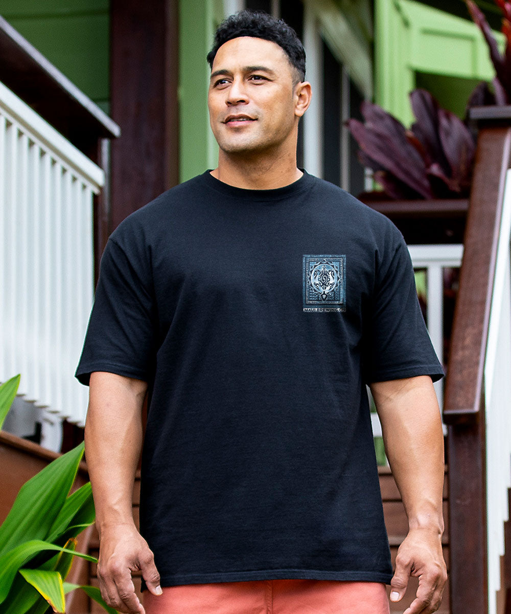 A man wearing a black crewneck t-shirt with a small square graphic on the left chest, standing outdoors in front of a house with green plants. View 3