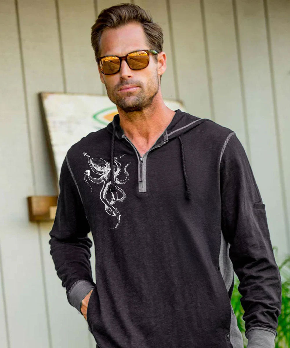 A man wearing a dark gray quarter-zip hoodie with a white snake graphic on the chest, sunglasses, and standing in front of a light-colored paneled wall.
