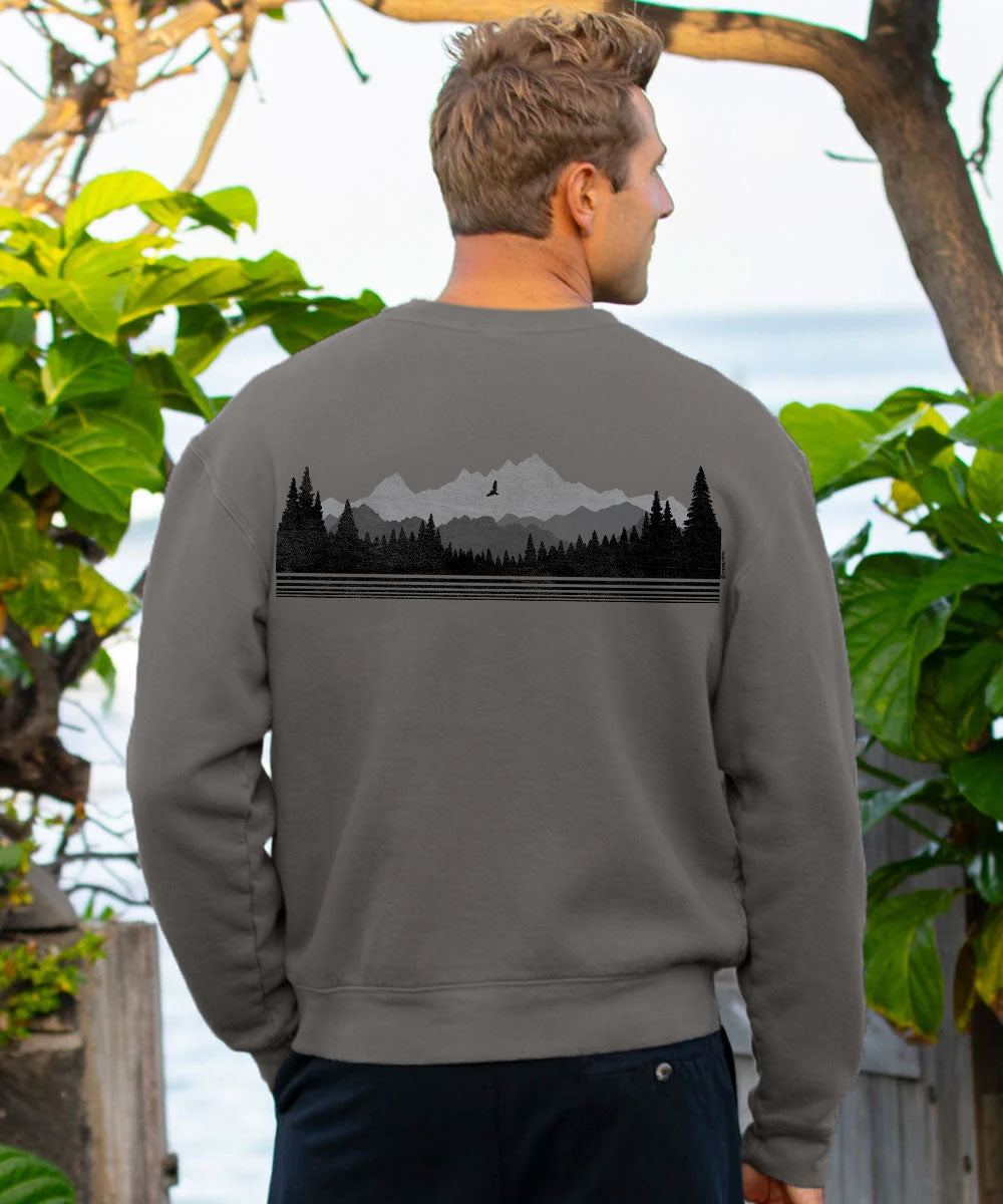 A man viewed from behind wearing a dark gray crewneck sweatshirt with a black mountain and pine forest graphic across the upper back, set against an outdoor background with green foliage and water. View 3
