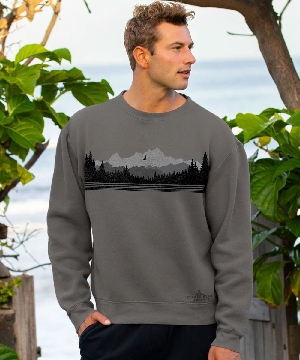 A man wearing a dark gray crewneck sweatshirt with a black and gray mountain and forest graphic, standing outdoors in front of a blurred background of green leaves and water. View 2