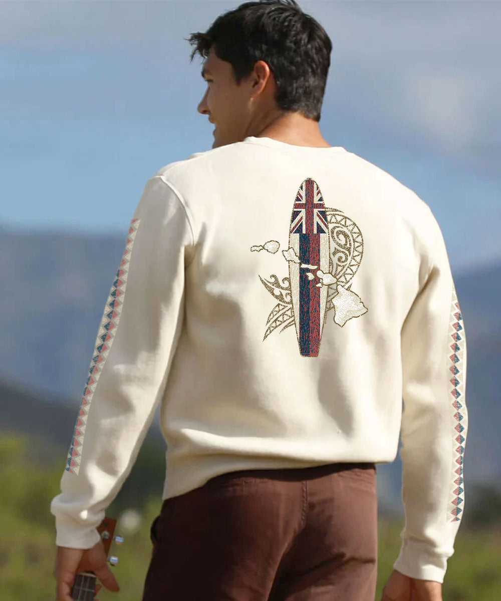 A man viewed from behind wearing a cream-colored crewneck sweatshirt with a detailed back graphic featuring a surfboard, Hawaiian islands, and Polynesian motifs, paired with brown pants in an outdoor setting. View 2
