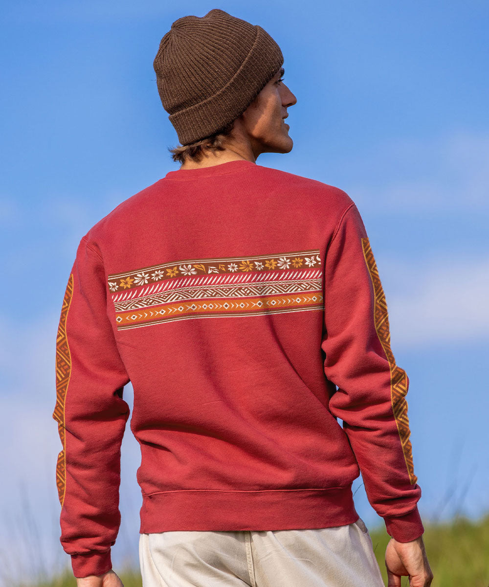A man wearing a brown knit beanie and a red crewneck sweatshirt with a decorative geometric pattern across the upper back and along the sleeves, photographed outdoors against a blue sky. View 3