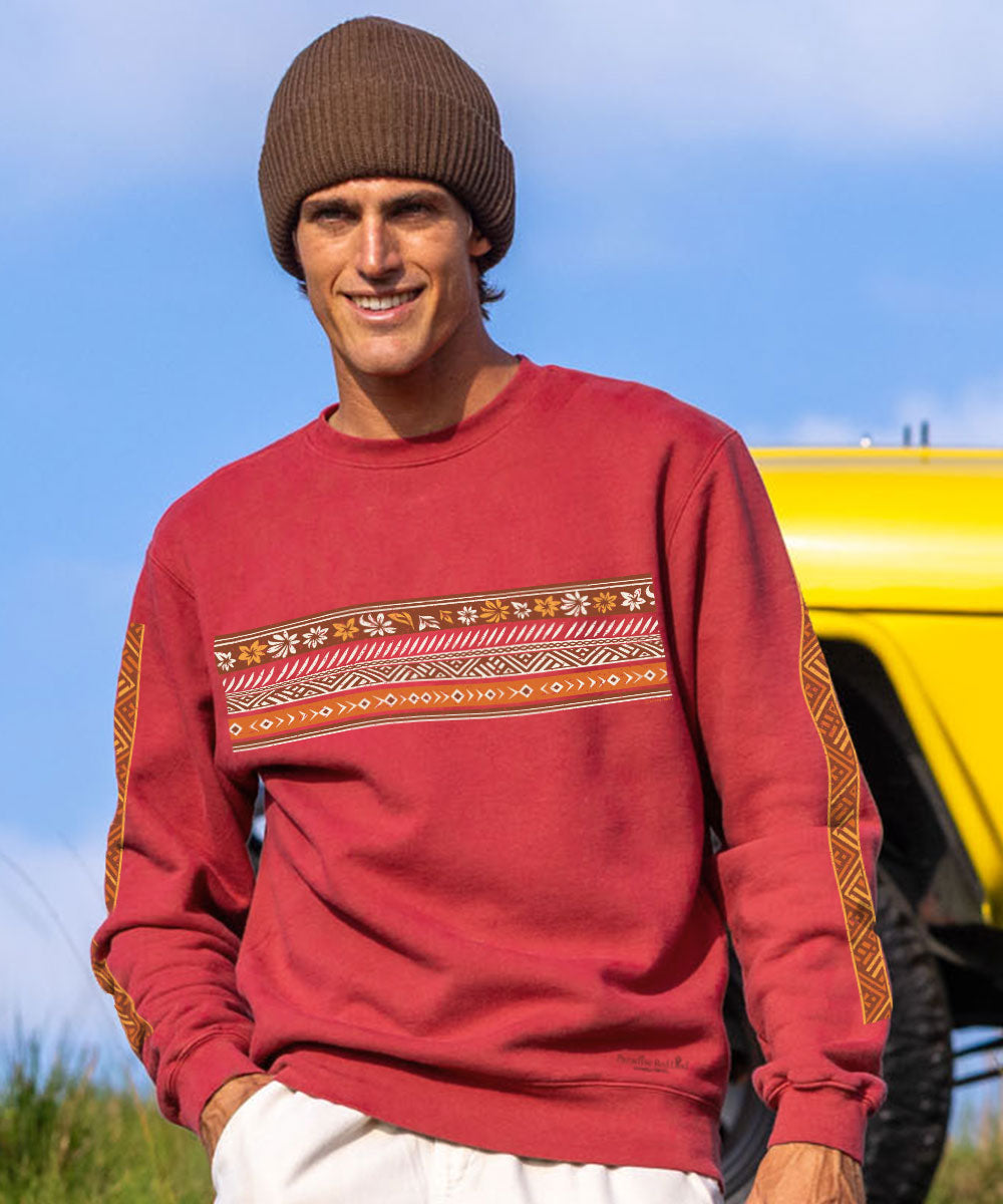 A man wearing a rust-red crewneck sweatshirt with a geometric patterned chest band and patterned sleeve cuffs, paired with a brown knit beanie and white pants, standing outdoors next to a yellow vehicle. View 2
