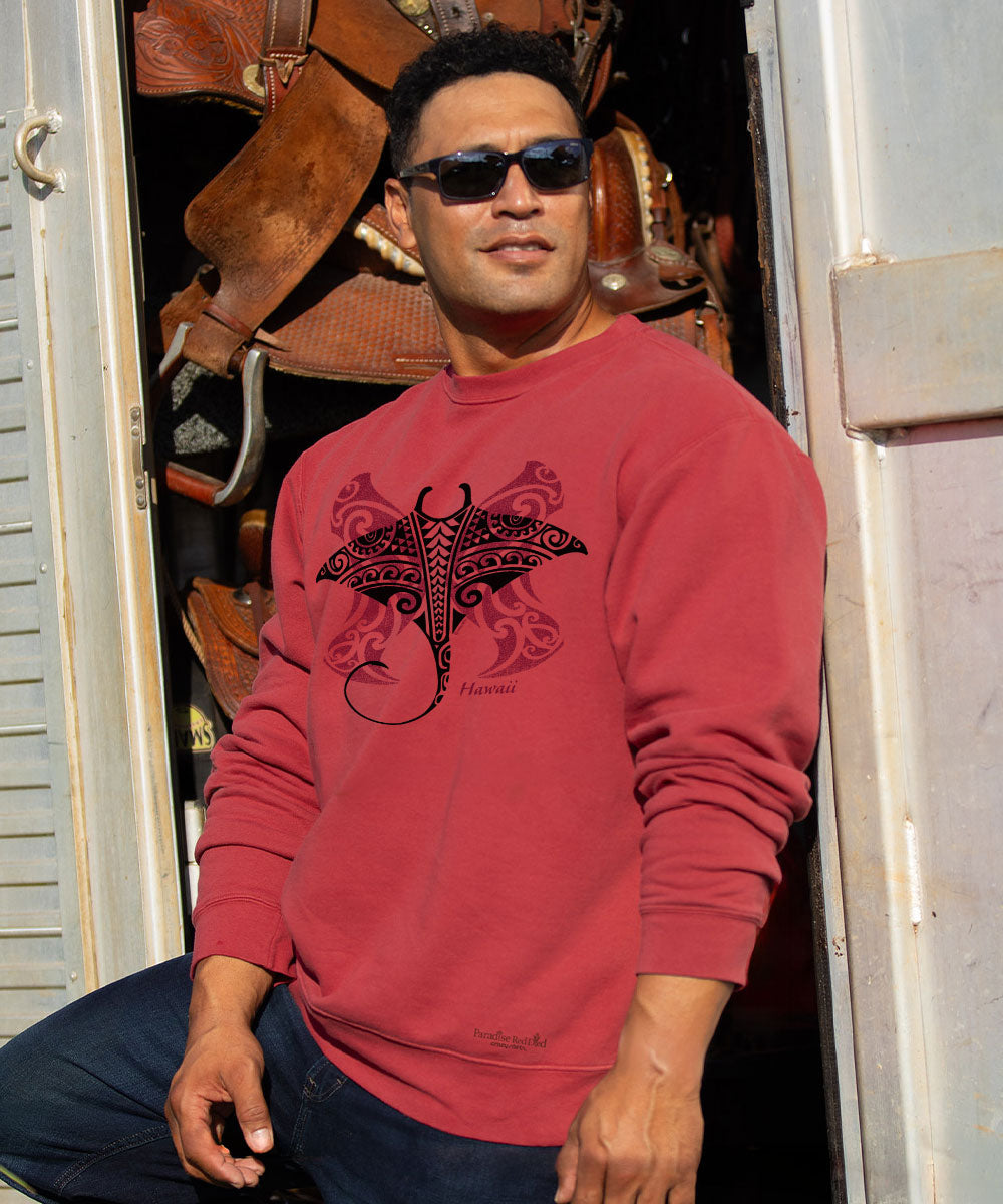 A man wearing a red crewneck sweatshirt with a black manta ray graphic and the brand name 'Hawaii' printed below it, standing in front of a doorway with leather gear in the background.