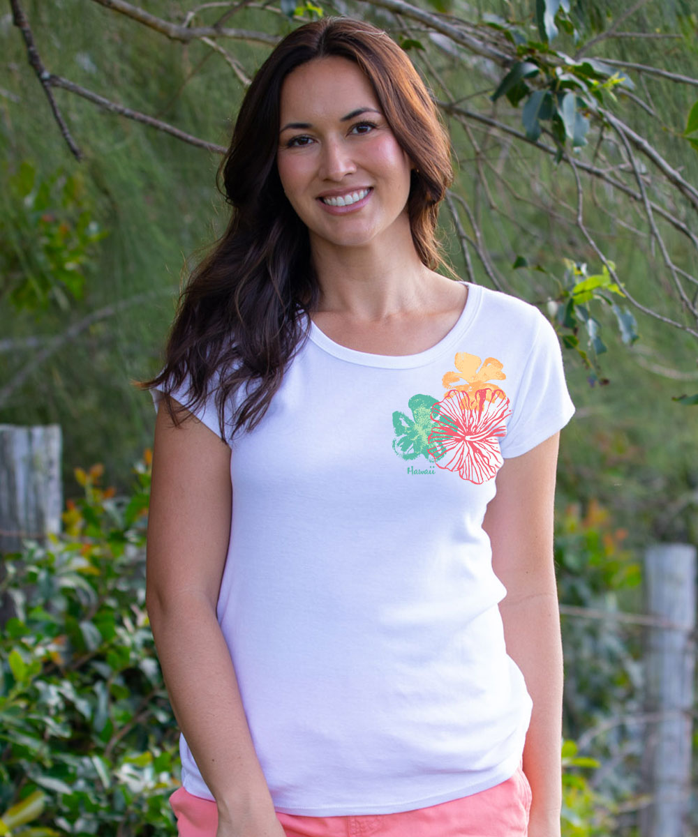 A woman wearing a white short-sleeve t-shirt with a colorful floral graphic on the left chest, standing outdoors in front of green foliage. View 2
