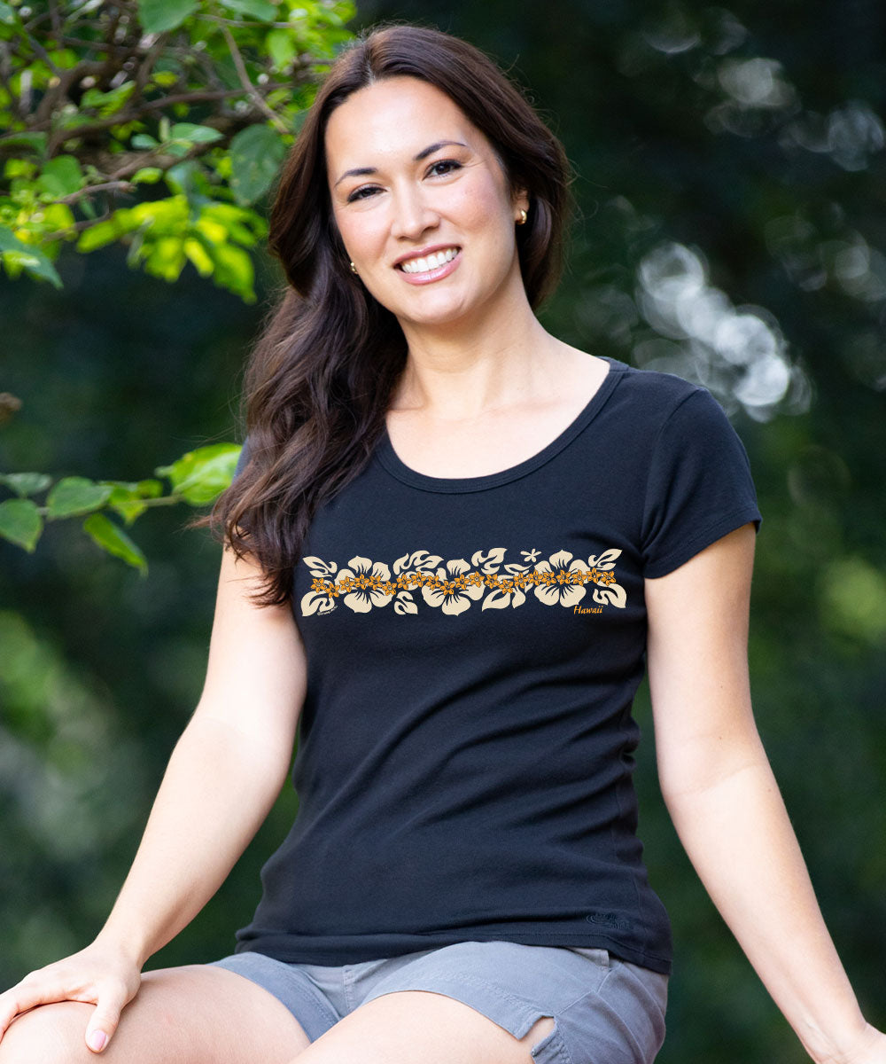 A woman wearing a black scoop-neck t-shirt with a horizontal gold and white floral pattern across the chest, posed outdoors against a soft green leafy background. View 2