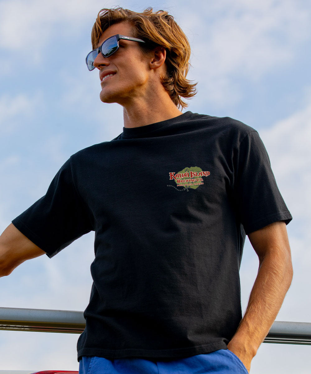 A man wearing a black short-sleeve t-shirt with a small 'Eagles Nest Brewing' logo on the left chest, photographed outdoors against a cloudy blue sky. View 3