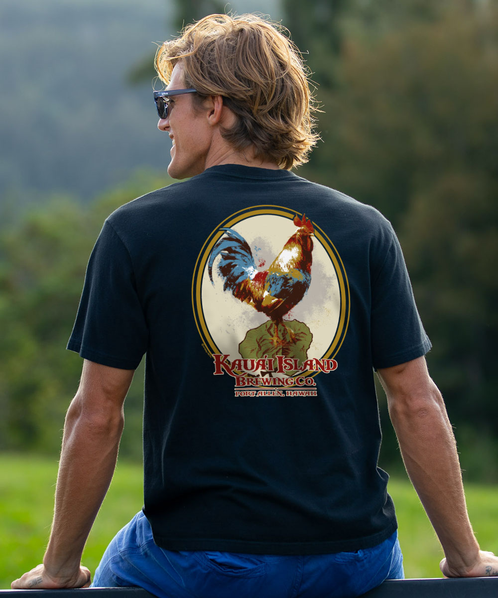 A man wearing a black t-shirt with a large graphic of a rooster on the back, photographed outdoors against a blurred green background. View 2