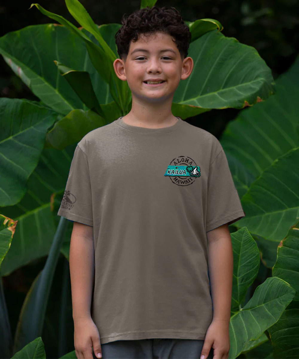 A young boy wearing a taupe brown short-sleeve t-shirt with a circular 'Kokoahua' logo on the left chest, standing in front of a lush background of large green tropical leaves. View 3