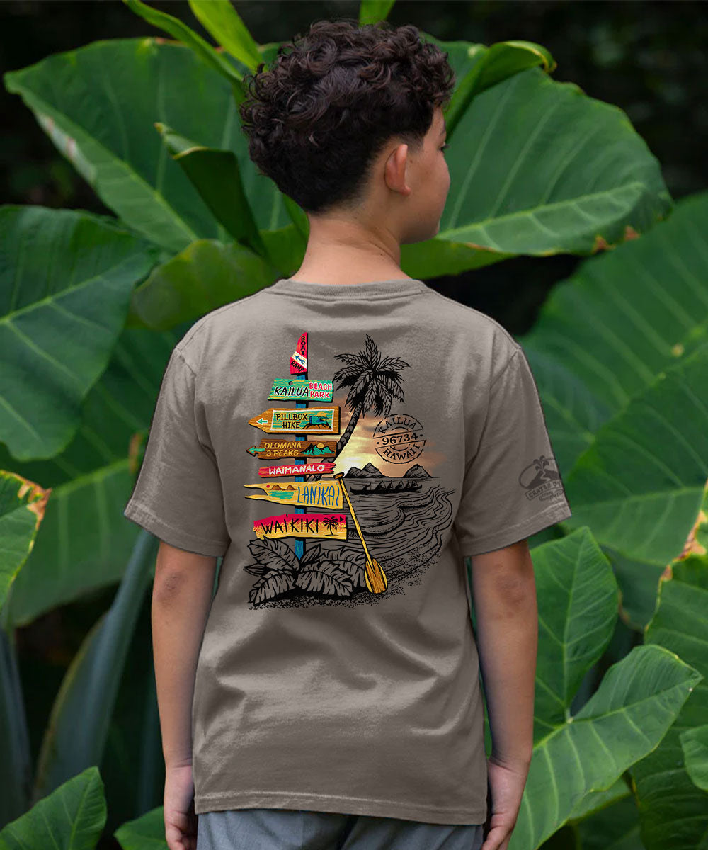 A child wearing a taupe brown t-shirt, viewed from behind, featuring a large multicolor graphic print of a tropical beach scene with a palm tree, sunset, and colorful surfboard signs. The background consists of lush green tropical leaves. View 2