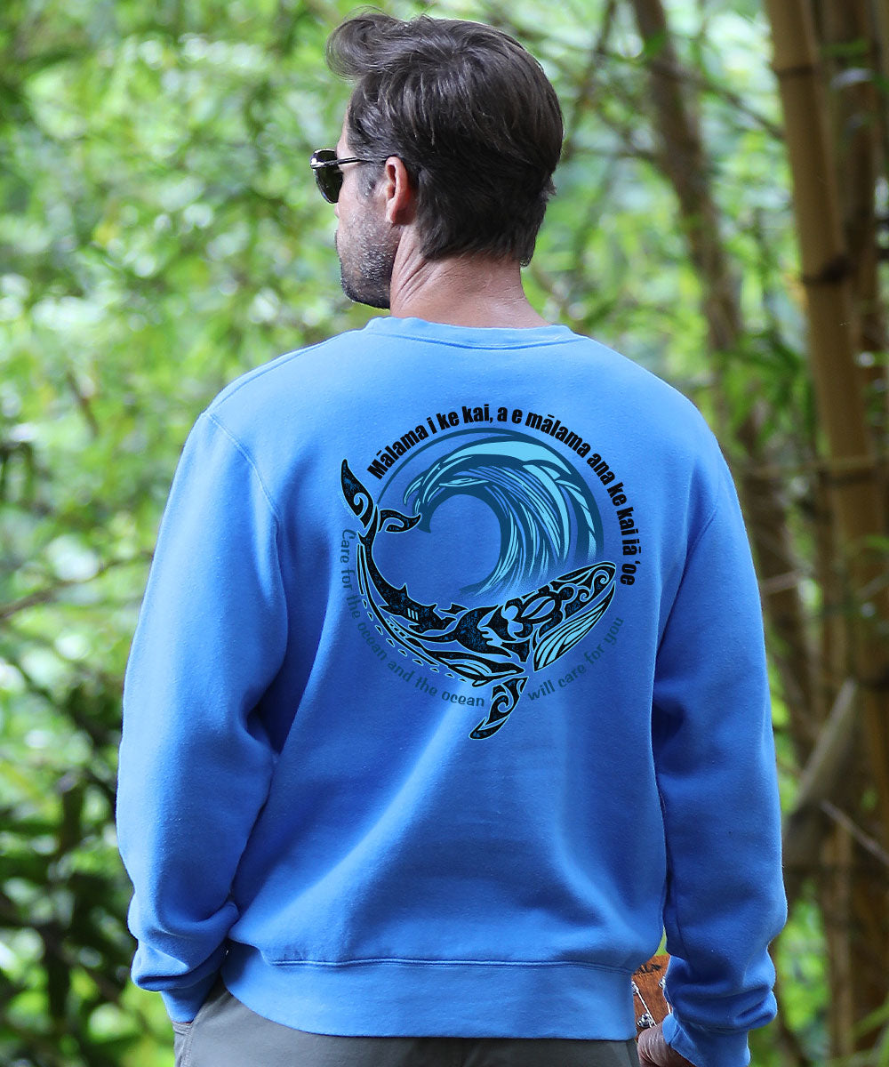 A man viewed from behind wearing a bright blue crewneck sweatshirt with a large Polynesian wave graphic, set against a blurred green forest background. View 2