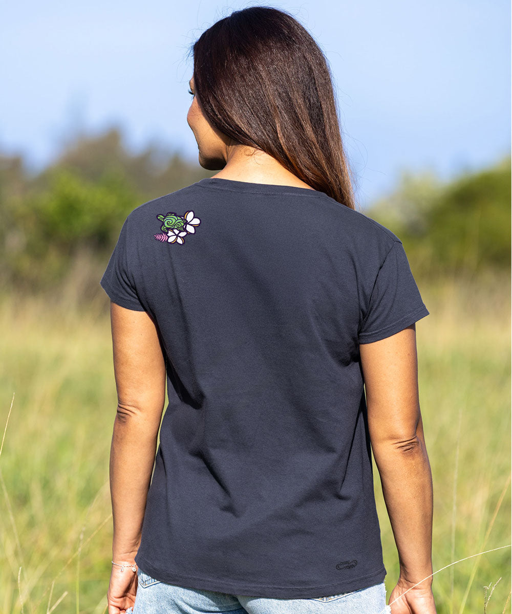 A woman viewed from behind wearing a dark charcoal gray t-shirt with a small green and white turtle graphic on the left shoulder, standing in an outdoor grassy field. View 3