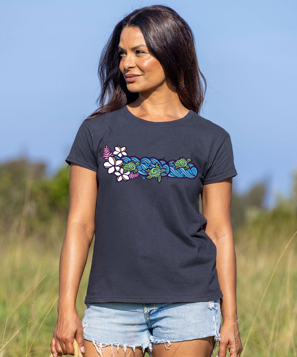 A woman wearing a dark charcoal gray t-shirt with a colorful graphic of green sea turtles and white flowers spelling 'Hawaii', paired with light blue denim shorts, standing in an outdoor grassy field under a blue sky. View 2