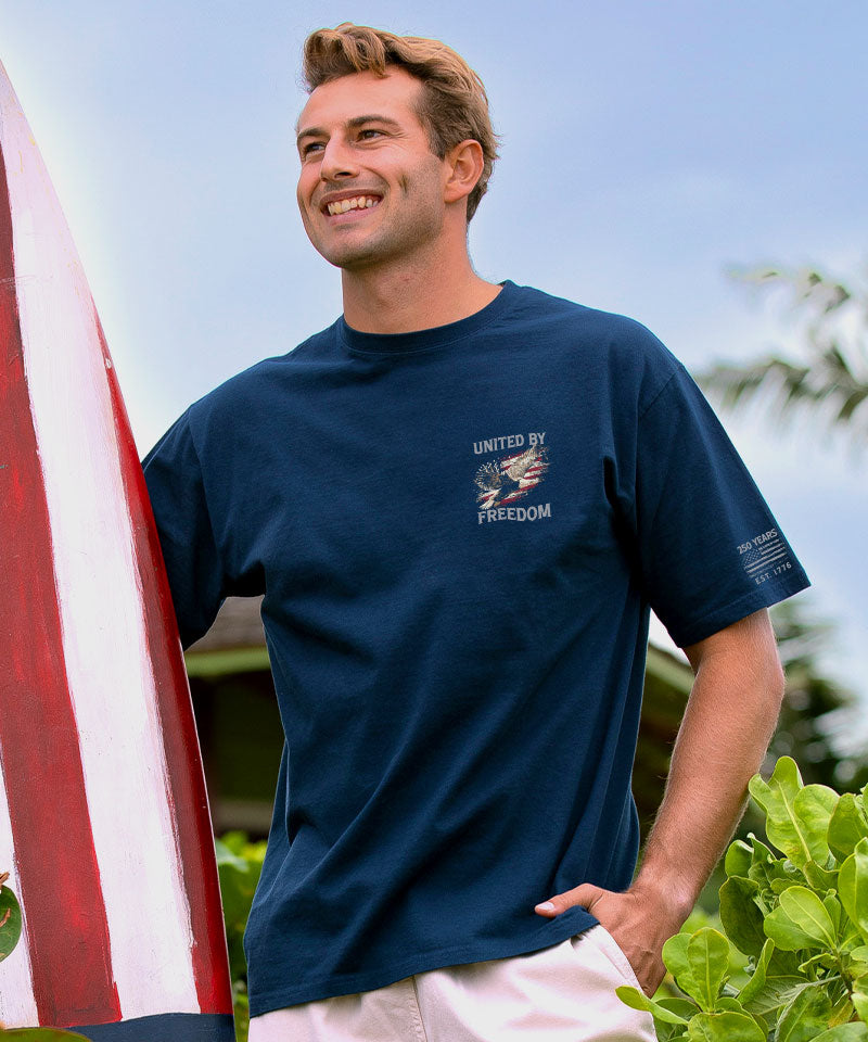 Man wearing a navy blue t-shirt with a logo, holding a surfboard outdoors. View 3