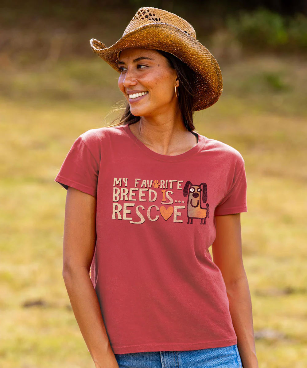 A woman wearing a rust-red graphic t-shirt and a straw cowboy hat, smiling outdoors in a grassy field. The t-shirt features the text 'MY FAVORITE BREED IS. View 2