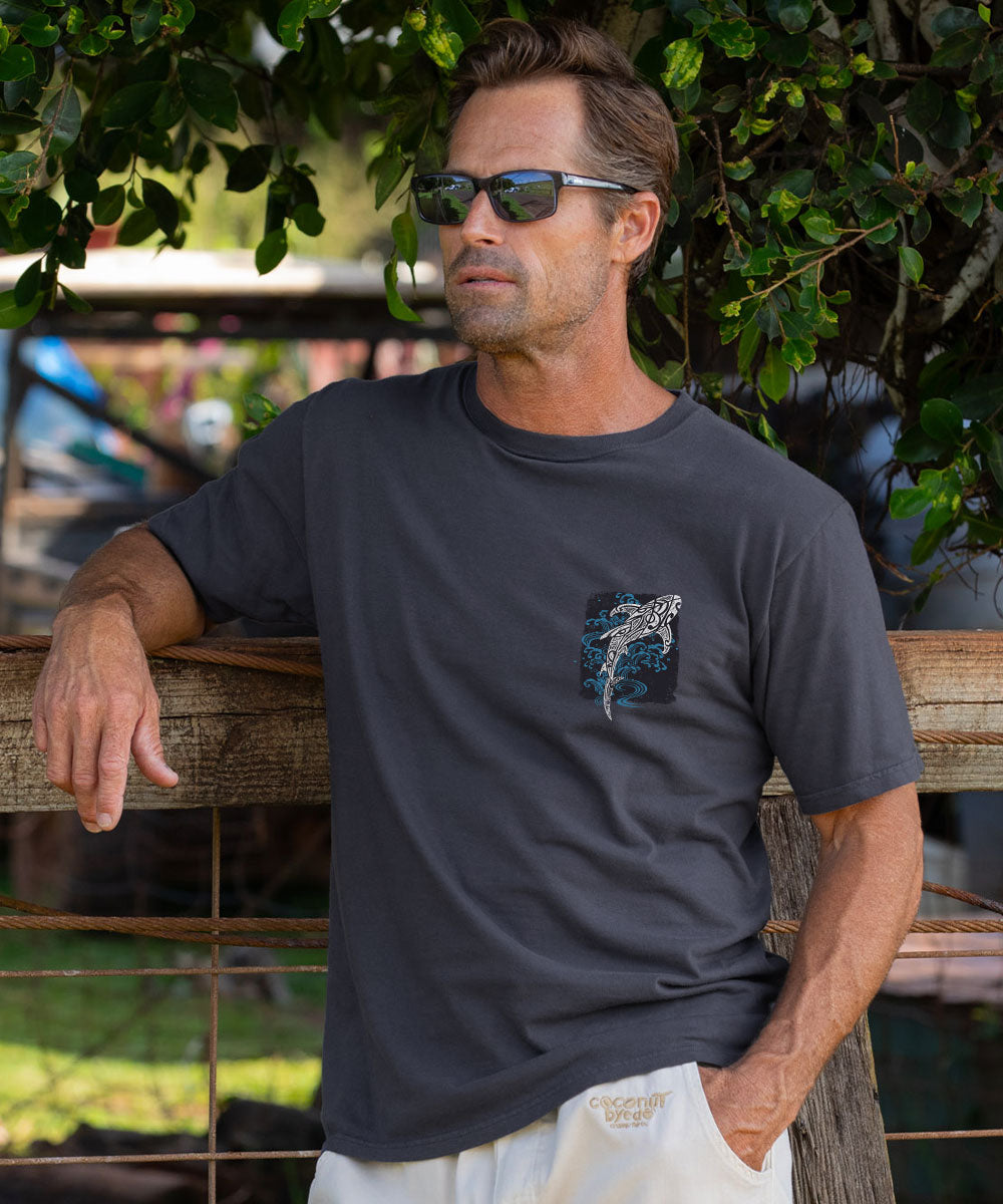 A man wearing a dark gray crewneck t-shirt with a shark graphic on the left chest, leaning against a wooden fence outdoors under green foliage. View 3