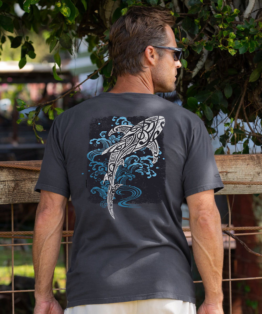 A man wearing a dark gray t-shirt with a large graphic of a stylized shark and blue waves on the back, photographed outdoors in front of a wooden fence and green foliage. View 2