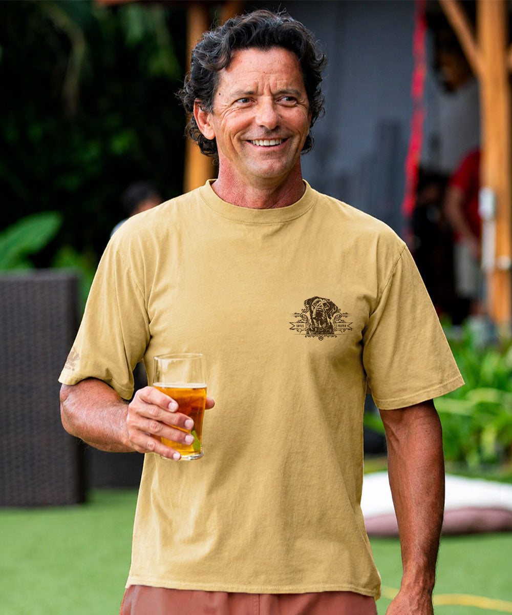 A smiling man wearing a tan graphic t-shirt with dog graphic, holding a glass of beer in an outdoor setting. View 3