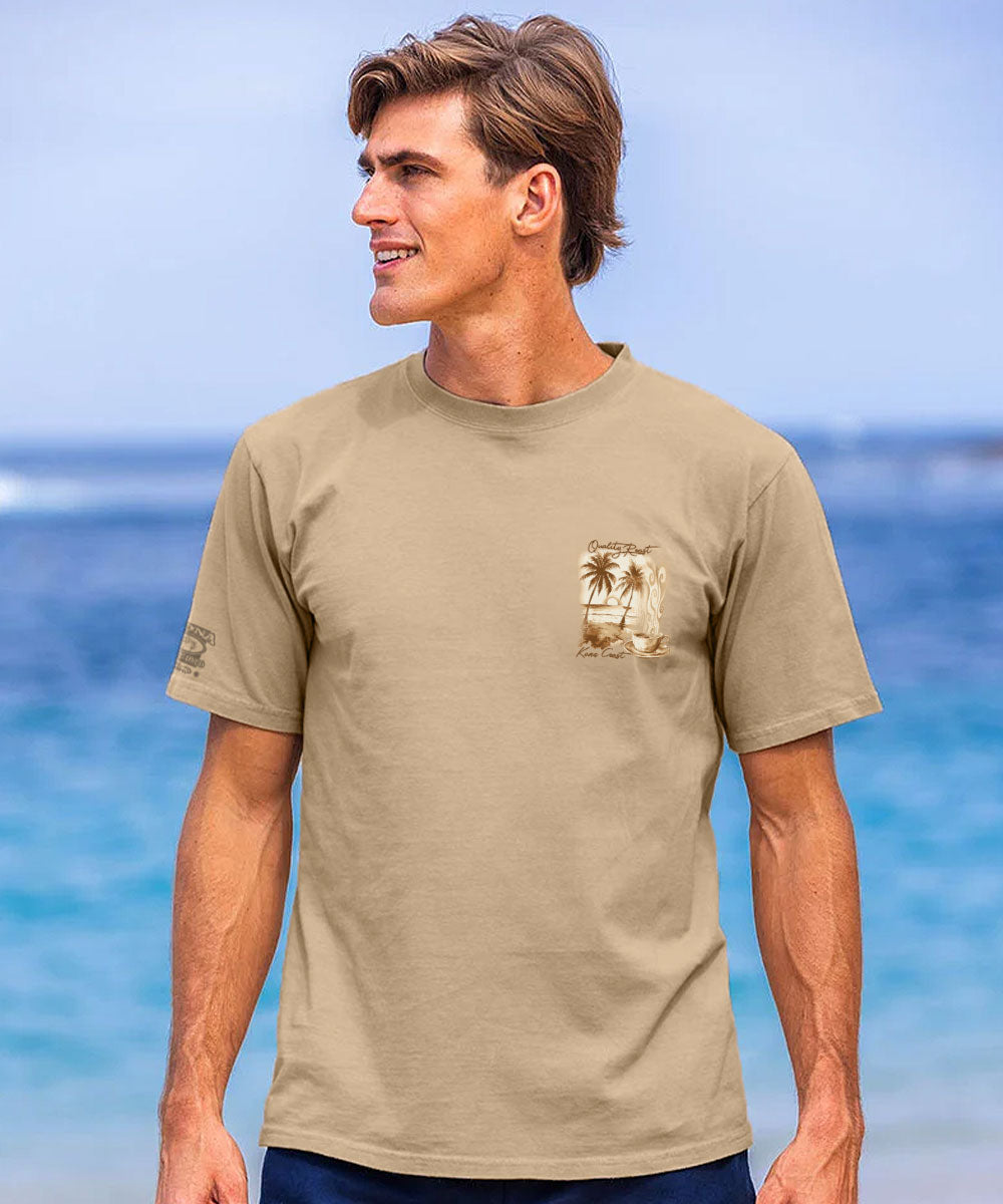 A man wearing a tan crewneck t-shirt with a small palm tree graphic on the left chest, standing against a blurred beach background. View 3