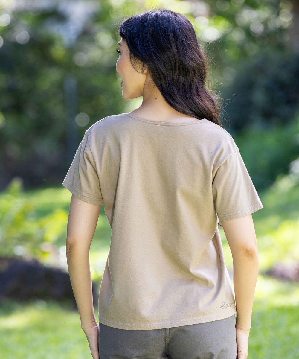 A woman viewed from behind wearing a light beige short-sleeve crewneck t-shirt and gray pants, standing outdoors in a sunlit grassy area with a blurred green background. View 3