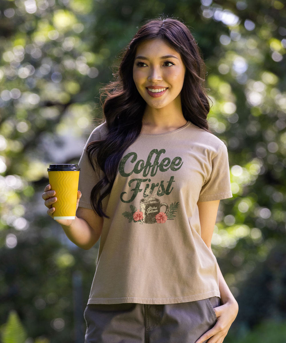 A woman wearing a beige graphic t-shirt with 'Coffee First' printed in green script with a mug and flowers, holding a yellow paper coffee cup against a blurred green outdoor background. View 2