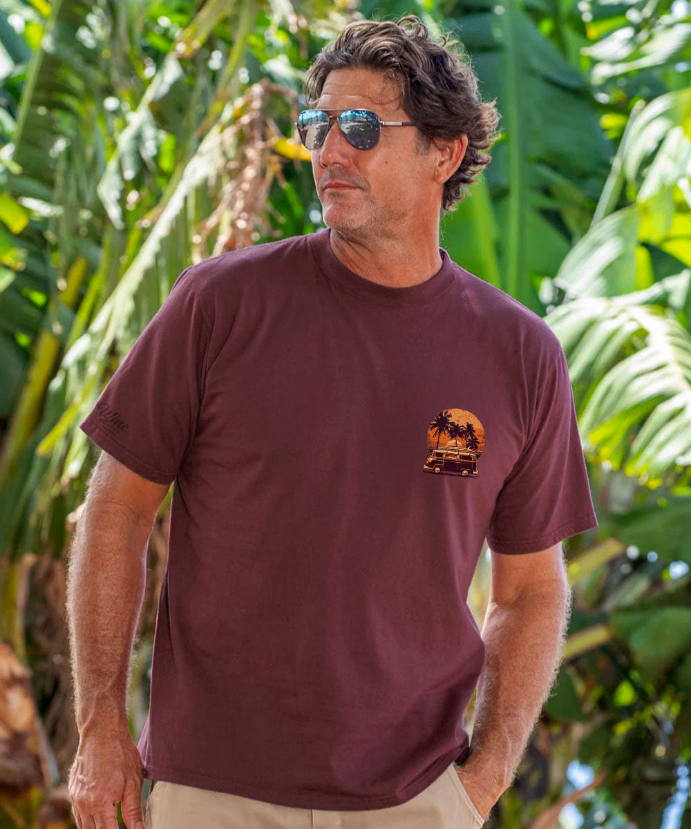A man wearing a short-sleeve crewneck t-shirt in maroon with a small orange and yellow graphic on the left chest, set against a background of lush green tropical leaves. View 3