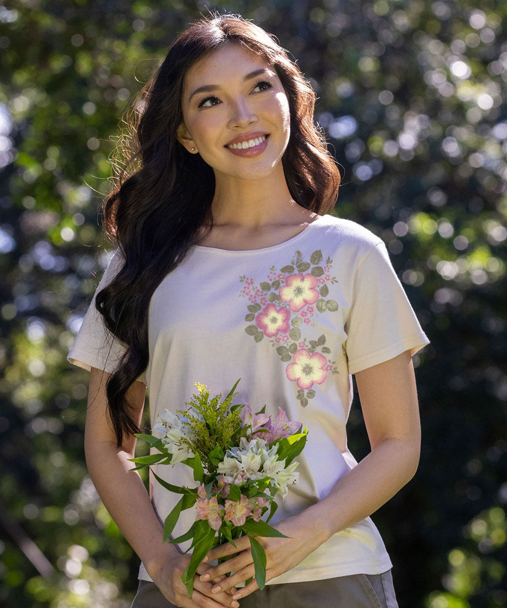 A woman outdoors wearing a cream-colored short-sleeve t-shirt with a pink and green floral graphic on the front, holding a small bouquet of white and pink flowers. View 2