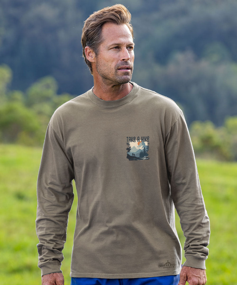 A man wearing a long-sleeve olive brown t-shirt with a 'Take a hike' graphic on the left chest, standing in a grassy field with a blurred forest background. View 3
