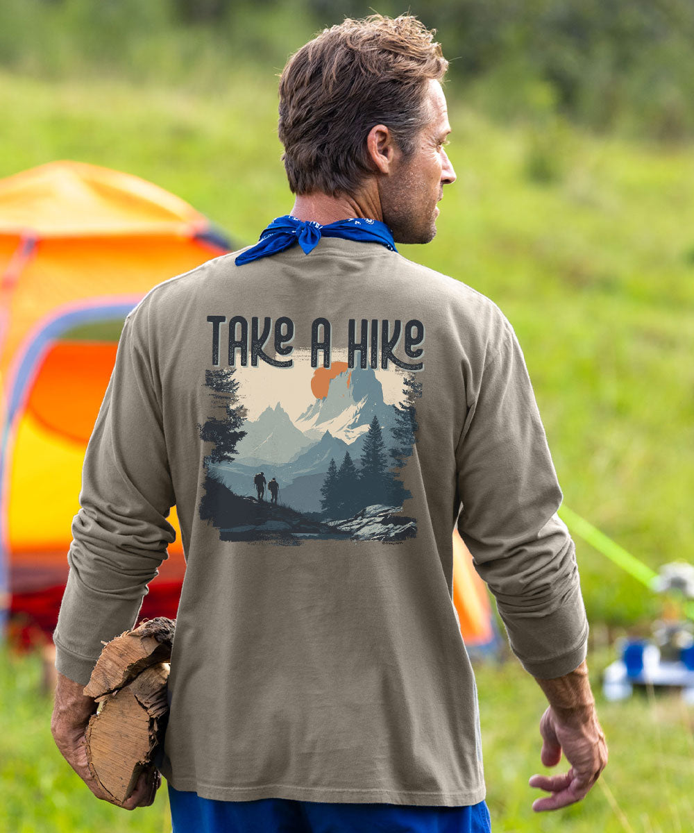 A man seen from behind wearing a long-sleeve olive green t-shirt with a 'TAKE A HIKE' graphic on the back, standing in a grassy outdoor camping setting with a tent in the background. View 2