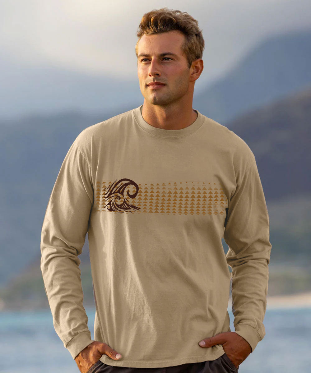 A man wearing a beige long-sleeve t-shirt with a brown wave graphic and a horizontal line of small geometric shapes across the chest, set against a blurred beach and mountain background.